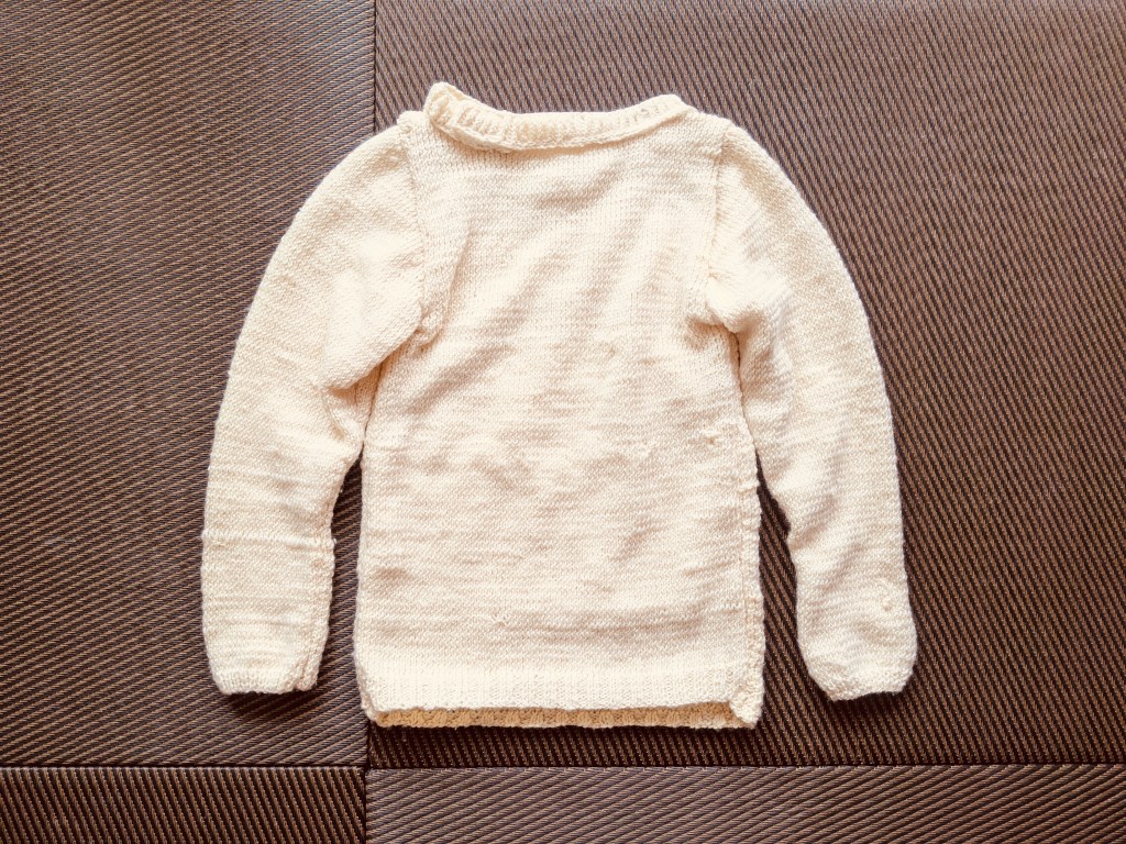 An off-white sweater lies spread out on the floor.
The collar is thick and uneven. The sleeves are oddly wide, while the body is visibly too narrow.
